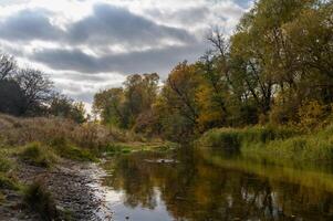 Autumn reflections along a serene river lined with vibrant trees under a cloudy sky photo