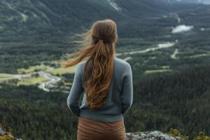 A woman with long hair standing on a mountain top looking at a valley photo