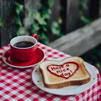 A red coffee cup sits on a checkered tablecloth alongside a piece of toast with Have a nice day written inside a heart made of jam. photo