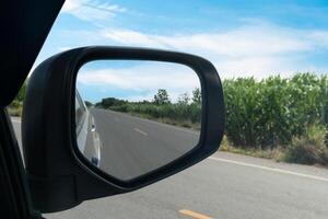 Inside view of mirrors wing. Rear view of a gray car with asphalt road and green trees in the daytime. photo
