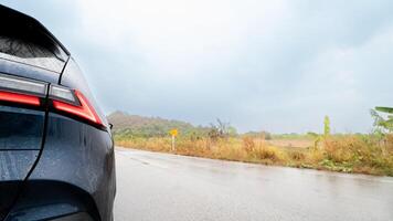 Car rear with a dark blue or black color and a red taillight detail. Car on a wet gray asphalt road. Indicating recent or ongoing rain. Background blurred landscape of fields and tree under a cloudy. photo