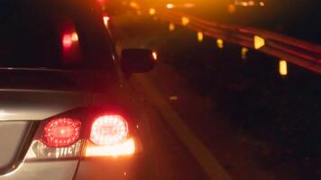 Rear of a car at night with illuminated tail lights and a side mirror visible. Scene is dark with a blurred background suggesting motion or low light conditions. photo
