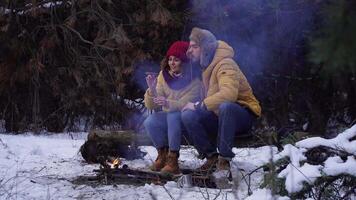 Young man and woman near a campfire in the forest in winter. Tourists travel together. The guys are smiling and chatting. Travel and adventure concept. video