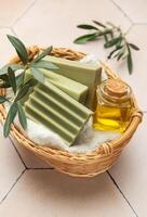 Olive soap, olive oil and branches resting in wicker basket on beige tile photo