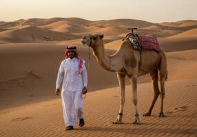A Saudi Gulf Arab man wearing a golden headband and a bisht standing next to a camel in the desert photo
