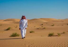 a Saudi Gulf Arab man wearing a ghutra and a bisht walking in the desert and looking at something, a desert background photo