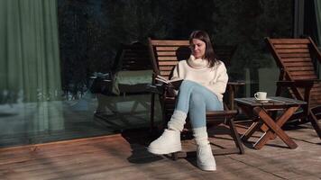 Comfortable woman enjoys reading a book while sitting on a wooden chair on a terrace during a sunny winter day, a cup of coffee on a small table beside her video