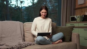 Young woman enjoys her leisure time, comfortably seated on a sofa in a cozy living room, browsing on a digital tablet against a backdrop of a tranquil evening scene visible through a large window video
