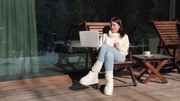 Remote working professional wearing white sweater and blue jeans, sitting on wooden chair at terrace, using laptop and enjoying hot beverage during winter morning video