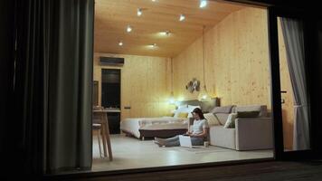 Wooden walls and warm lighting create cozy atmosphere as a young woman sits on the floor with a laptop, working remotely from a tiny house with a glass wall open to the night video