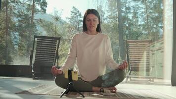 Young woman in sportswear practicing yoga and meditation with burning incense in front of her smartphone placed on a tripod in a modern house with large windows and mountain view video