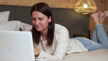 Female professional relaxing on bed, using laptop while enjoying comfortable workspace with wooden walls and stylish wicker lamp illuminating cozy bedroom environment video