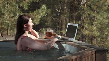 Professional freelancer working remotely from luxurious hot tub, surrounded by serene forest landscape, using laptop while enjoying tea and embracing digital nomad lifestyle video