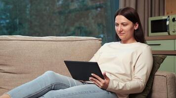 Comfortable woman using digital tablet while relaxing on sofa in living room, enjoying online content, social media, or working remotely from home video