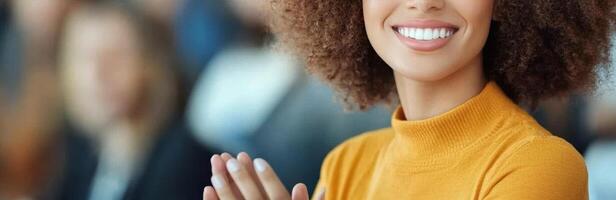 smiling woman with curly hair claps her hands in modern conference setting, expressing joy and engagement. Her bright orange sweater adds warmth to scene photo