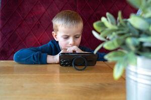 A 4-5-year-old European boy sits at the table, watching cartoons on his phone, leaning in closely. Immersed in digital entertainment, he focuses on the screen, enjoying his favorite animated content. photo