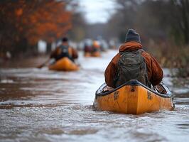 Kayakers navigate flooded road, autumn trees in background, disaster relief photo