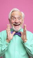 Excited senior man showing enthusiasm and surprise on pink background. Elderly man with grey hair and beard wearing bow tie expressing excitement and surprise with raised hands photo
