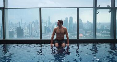Man chilling and relaxing in luxury pool on roof of skyscraper. Young 30s business male sitting on the edge of the roof top swimming pool in luxury hotel on background big city video