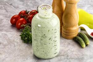 A bottle of dressing next to a pepper, tomato and cucumber photo
