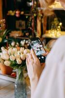 Close-up of person photographing bouquet of white tulip using smartphone in floral arrangement shop. Background features beautiful lighting and floral decor, creating warm and inviting atmosphere photo