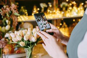 Close-up of person photographing bouquet of white tulips using smartphone in floral arrangement shop. Background features beautiful lighting and floral decor, creating warm and inviting atmosphere photo