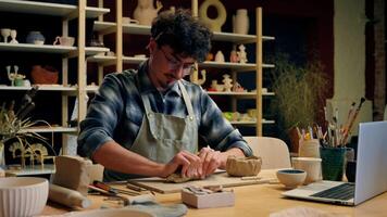 Caucasian man guy male potter designer artist art sculptor modeling clay handmade with rolling pin on table pottery workshop using laptop tutorial handcrafting ceramics crockery design hobby video