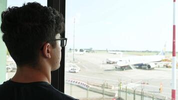 Caselle Airport, Turin, Italy. August 13, 2024. A young Caucasian man looks out the window at the runway and takes a photo with his cell phone. Travel lifestyle and use of technology. video
