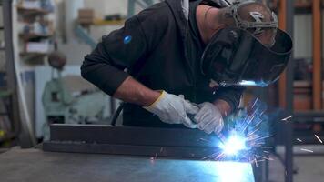 Performing a weld, the man in the welding mask is positioned with the welding machine on the welding point. When he activates it, a blue glow with smoke and orange sparks illuminates the scene video