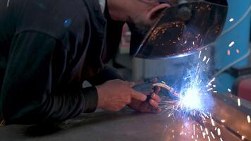 Performing a weld, the man in the welding mask is positioned with the welding machine on the welding point. When he activates it, a blue glow with smoke and sparks illuminates the scene video