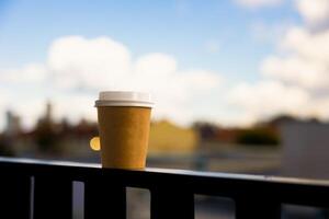 A paper cup for coffee with white lid stands on the railing on sunny summer day photo