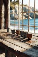 Morning light floods a rustic wooden table with five ceramic mugs overlooking a serene coastal view photo