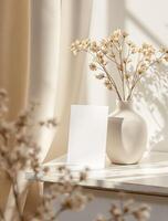 A white vase with dried flowers and a blank card photo
