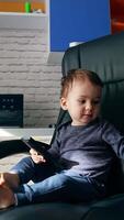 Beautiful baby in the office chair with remote control in hands. Kid turns to the table with laptop and puts the remote control there. Vertical video