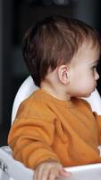 Cute dark-haired little boy sits at a feeding table with bagels in front of him. Mom gives the full spoon to her son and he slowly opens the mouth. Blurred backdrop. Vertical video