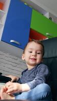Smiling cute kid sitting in the turning black leather chair. Toddler turns to the table to take remote control. Low angle view. Vertical video