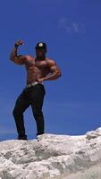 Low angle view of the strong fit bodybuilder showing his biceps. Handsome athletic man showing his muscles while standing at the sand with blue sky at the background. Vertical video