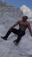 Man with perfect body pumping up muscles and enjoying of the summer. Multiracial man sitting at the sand. Grey rocks background. Vertical video