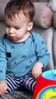 Dark-haired Caucasian toddler boy sitting on the floor next to the sofa. Adorable child plays with bright toys in front of him. Vertical video