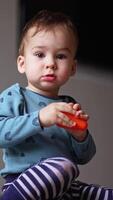 Baby boy sitting on the floor among the toys stares at camera. Toddler plays with a plastic toy squeezing it in hands. Low angle view. Vertical video