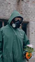 Survivor holding flowers with ruined building background. Person in chemical safety suit and respirator standing with flowers in hands in abandoned place and stretching t to the camera. Vertical video