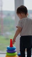 Toddler boy comes up to a colorful pyramid near the panoramic window. Active happy kid assembles the toy. Vertical video