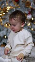 Little lovely boy in white clothes sits on the floor. Baby throws the red ball in front of him. Christmas tree at backdrop. Vertical video