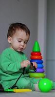 Calm Caucasian child sits on the floor playing with a black cord. Balls, pyramid and big yellow toy box near the kid. Vertical video