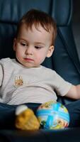 Beautiful Caucasian baby sits in a leather chair with a ball. Boy sits peacefully then takes a ball and stands up. Vertical video