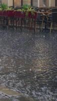 stream of water are flowing through the pavement in rainy weather. Raindrops is falling on the road against the background of the summer terrace of the restaurant. Vertical video