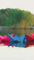 several multi-colored origami in the shape of ships are floating on the water in the lake in the evening on the background of the summer landscape. Vertical video