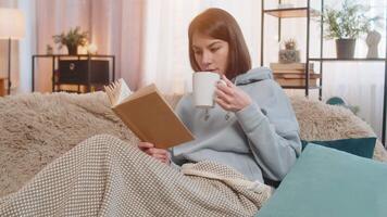 Smiling young woman reading interesting book while having cup of hot chocolate on couch at home video