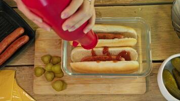 Two traditional hot dogs, made with meat sausage, are poured with spicy ketchup on a rustic kitchen table during cooking. video