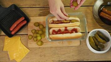 In the rustic-style kitchen, the chef prepares a traditional hot dog and adds slices of pickled cucumber to sausage rolls. Top view. video